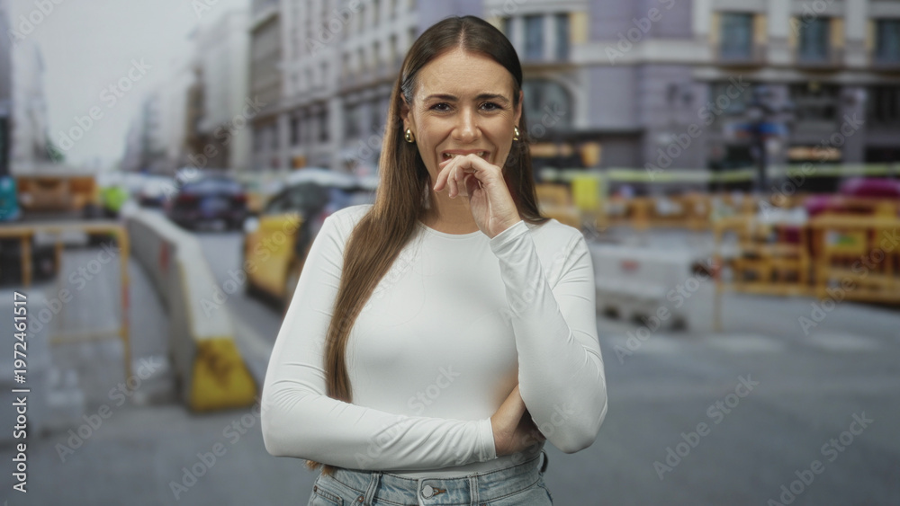 Fototapeta premium Young woman in white long sleeve top, hand near mouth in a shy laugh and crossed arms, standing on a busy city street with yellow taxis and construction barriers visible; playful confidence.