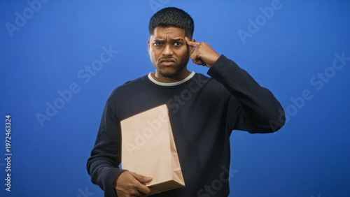 Fotografie Man holding a paper bag with finger to temple and furrowed brow in studio, facing an apparent delivery mixup and order delay; frustration