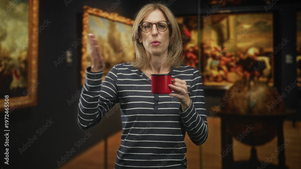 Obraz premium Woman holding red mug and touching chest while tasting coffee in museum, wearing glasses and striped shirt, surrounded by framed paintings and a globe; discomfort.