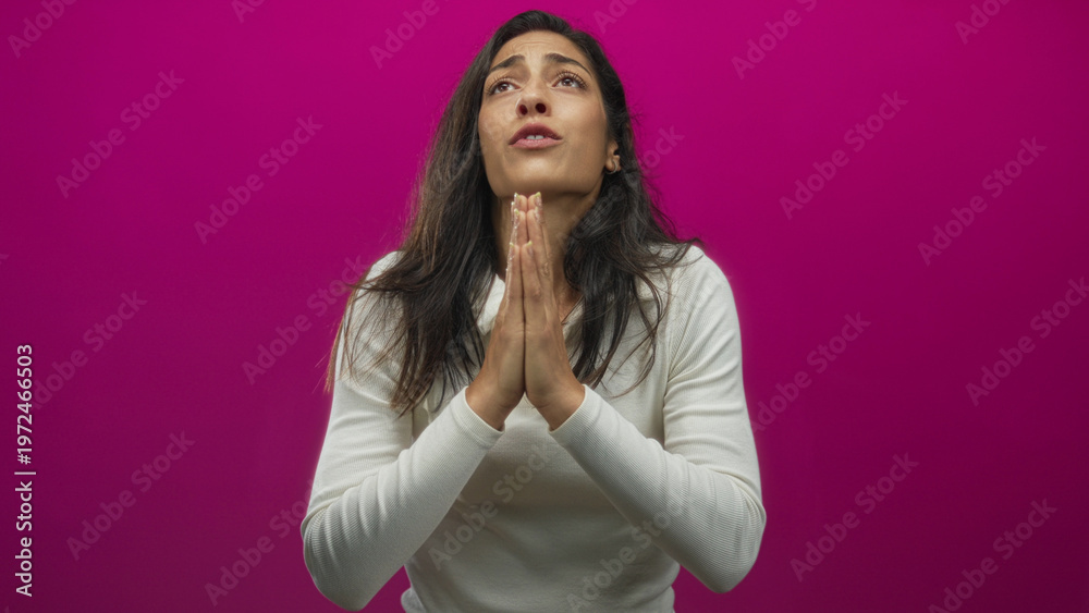 Obraz premium Young hispanic woman praying with clasped hands in studio against magenta backdrop; hopeful plea for comfort.
