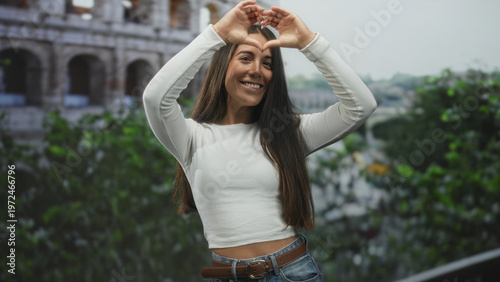 Photography Young woman making finger heart with right hand near chest, smiling in front of the colosseum building; travel joy