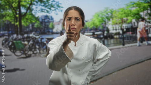 Woman chef pointing finger forward on a busy street by a canal with bicycles and buildings, stern expression; firm authority.