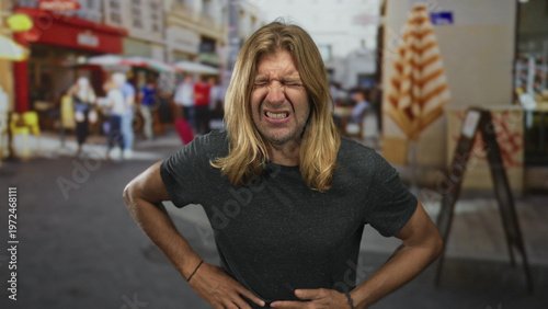 Schilderij op canvas Blond man with long hair visibly grimaces as he clutches stomach on street; pain discomfort nausea distress