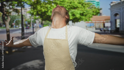 Canvas Print Man wearing an apron spreads out arms and tilts his head back under bright sunlight on a city street; serenity
