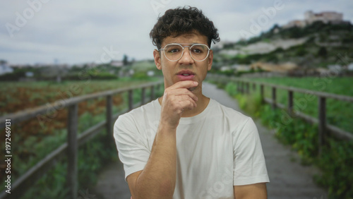 Cuadro en lienzo Hispanic man wearing glasses and a white shirt with hand on chin stands on a street by green fields; thinking thought pondering concern