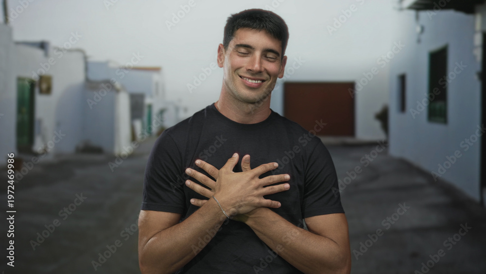 Fototapeta premium Smiling hispanic man wearing black shirt holds hands to chest on historic stone street in old town; self love gratitude.
