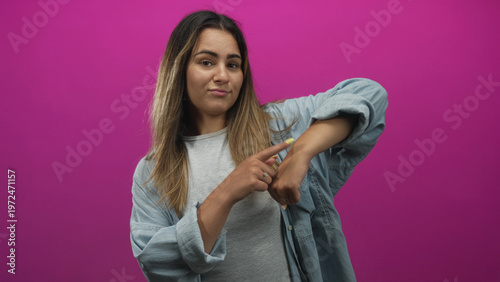 Photography Hispanic woman pointing finger to wrist and tapping wrist in pink studio wearing denim shirt and gray tee, long hair visible; impatience