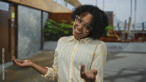 Photography Young african american woman with glasses holds palms up at building entrance in sunlight; warm welcome joy