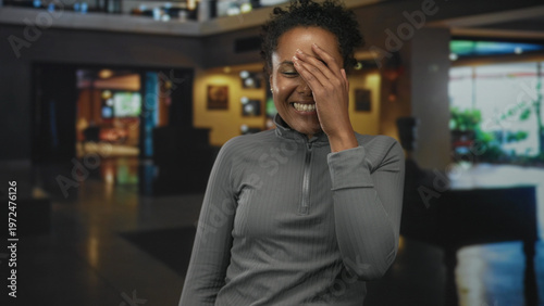 Canvas-taulu Young african american woman laughing and covering her face with her hand in a hotel lobby near seating area and reception desk; bashful happiness