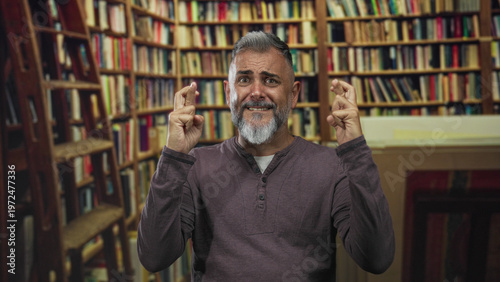 Принти на полотні Man crossing fingers and grimacing in a library surrounded by bookshelves and a ladder, facing camera and seated; hopeful anticipation