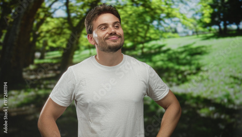 Young hispanic man smiling with hands on hips wearing white t shirt in green park outdoors under trees on a sunny day; confidence.