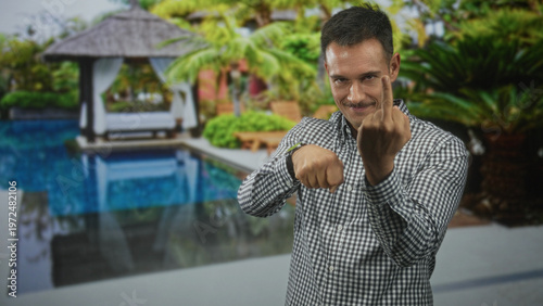 Slika na platnu Young man with moustache points and shows middle finger toward camera by poolside building; defiance