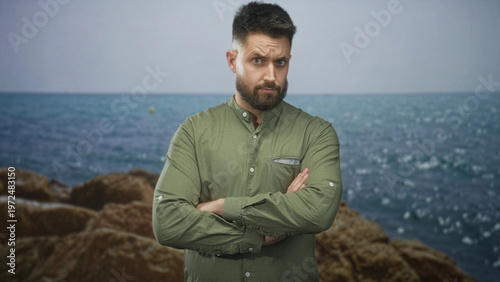 Billede på lærred Man with beard crossing arms, wearing green shirt, standing before rocky sea and boulders in studio; thoughtful resolve
