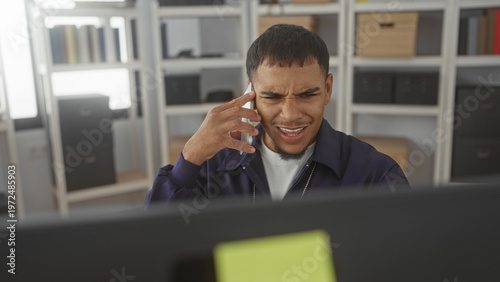 Tableau sur toile Man holding phone to ear in office, focused on monitor with furrowed brow and hand at temple; concern