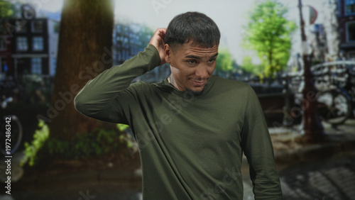 Εκτύπωση καμβά Man wearing green long sleeve shirt scratching head with hand and wry smile on cobbled street by canal and bikes, face and hand visible; awkward charm