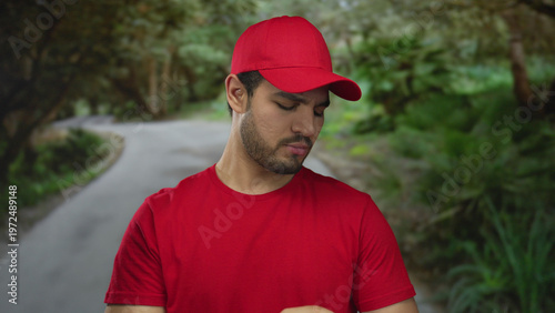 Photography Young hispanic man wearing red shirt and matching cap pointing finger amid lush green forest; determination focus clarity direction