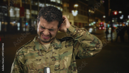 Fotografie Man wearing camouflage uniform and dogtags grimaces on busy city street at night under glowing streetlights; weariness