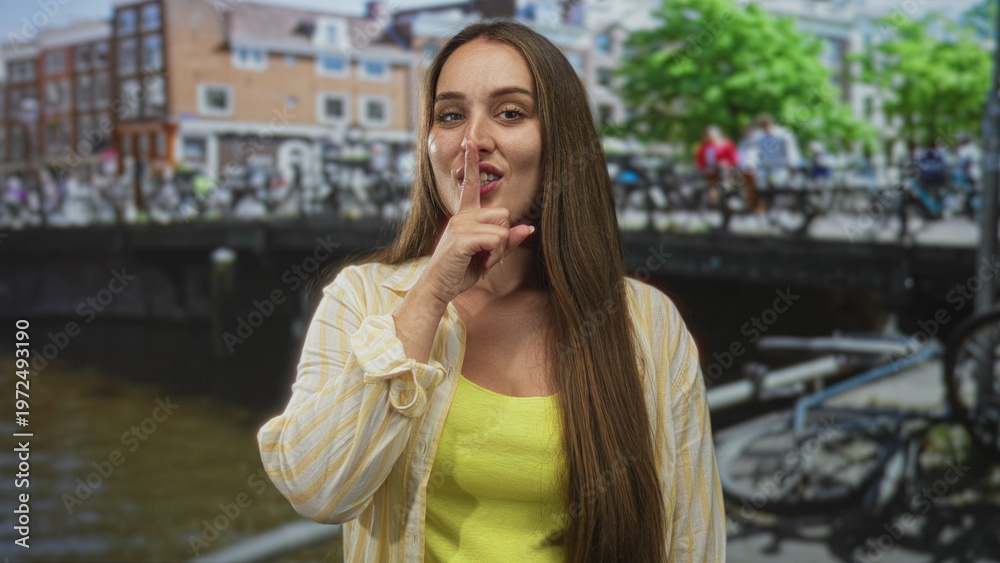 Fototapeta premium Woman putting finger to lips for silence on amsterdam canal street with bicycles and brick buildings; secret hush.