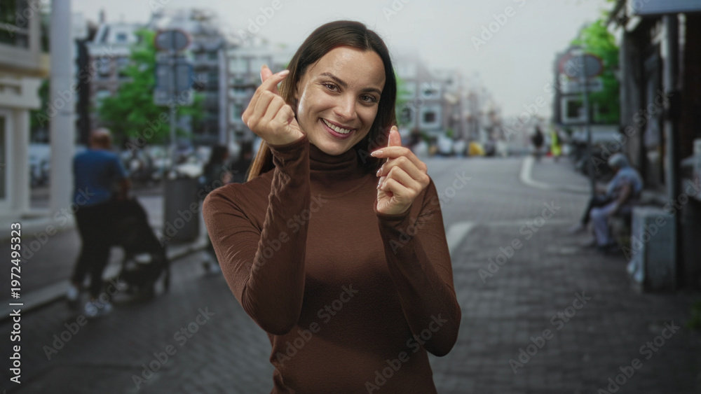 Fototapeta premium Woman smiling and making finger heart gesture with hands near chest in a brown turtleneck on a busy cobblestone street in city center; joy warmth.