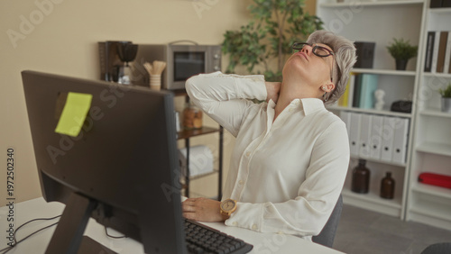 Woman in white shirt wearing glasses holding neck leaning back at office desk computer monitor; fatigue.