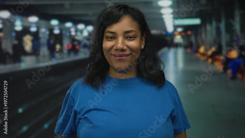 Canvas Print Woman in blue shirt standing on platform at train station under overhead lights smiling gently; quiet confidence