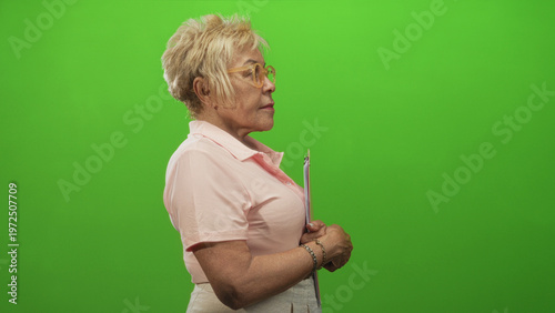 Senior woman in pink polo holds clipboard with hands clasped in profile at studio green screen; calm focus.