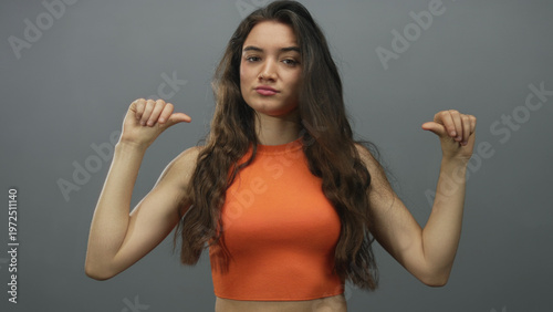 Bild auf Leinwand Hispanic young woman in orange crop top points thumbs to chest in grey studio; confidence self esteem empowerment