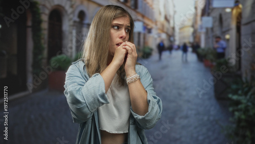 Tableau sur toile Young blonde woman covering mouth with both hands while wearing light blue denim jacket and bracelets on cobblestone street; anxiety