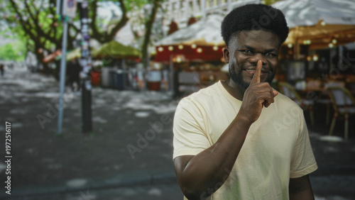 Tableau sur toile Young man smiling points finger and finger to lips for silence while standing near outdoor cafe on a city street; playful secrecy