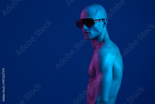 Bald man posing confidently in blue lighting with stylish sunglasses, showcasing a strong physique in a vibrant atmosphere. Perfect for fashion and advertising needs.