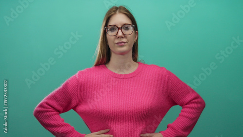 Fotografija Woman wearing glasses and pink sweater pointing finger forward with serious expression in teal studio setting; assertion