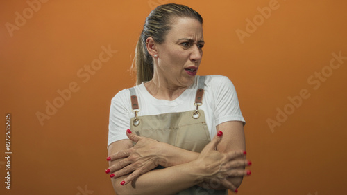 Photos Blonde woman wearing a white t shirt and overalls crossing arms across chest in studio with orange background; disgust