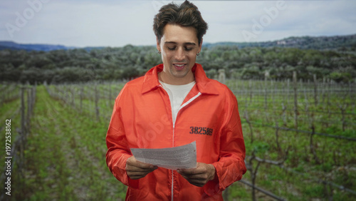 Man prisoner in orange jumpsuit reads a paper letter, holding it with both hands, pensive expression while standing among vineyard rows in a forest setting; remorse solitude.