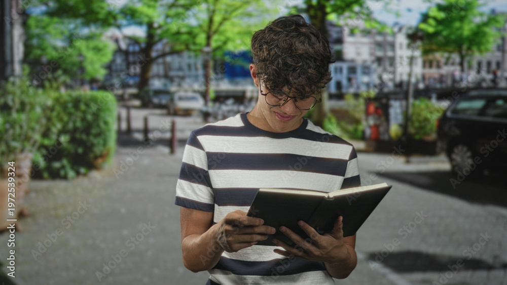 Obraz premium Teen boy in striped shirt holding open book with hands, reading on a tree lined urban street near parked cars; quiet concentration.