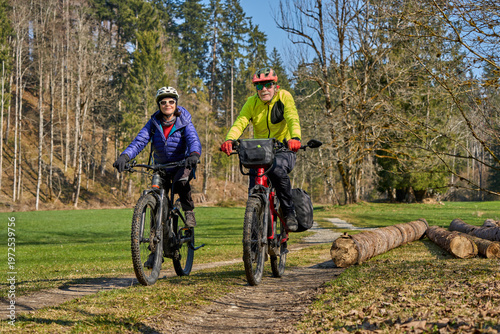 Happy senior couple enjoying an active lifestyle, riding electric bikes on a forest trail in spring. Outdoor recreation, healthy aging, and leisure time in nature on a sunny day.