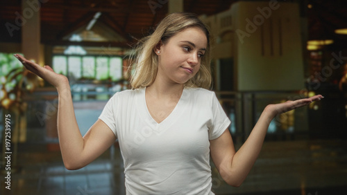 Obraz na plátne Woman in white tshirt standing palms up shrug at hotel lobby reception area, casual pose and neutral expression; indifference doubt ambivalence