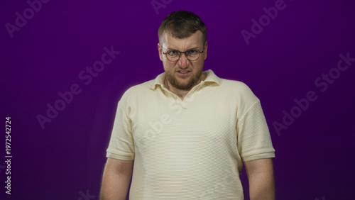 Bild auf Leinwand Man with glasses grimacing and squinting in studio against purple backdrop wearing cream polo shirt, hands down by sides; disgust discomfort