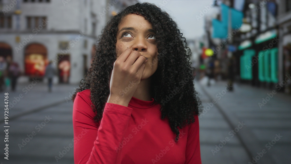 Fototapeta premium Woman in red top raises hand with open palm on street in front of building facade and smiles broadly with curly hair flowing; astonishment.