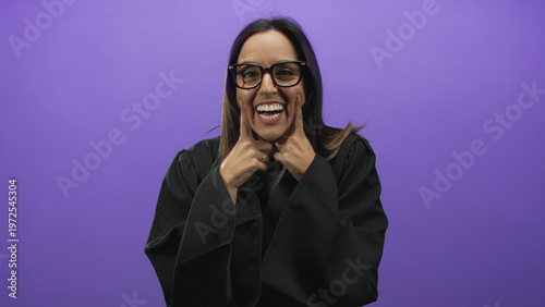 Фотография Woman judge wearing black robe and glasses presses cheeks with index fingers and puffs lips in a purple studio; playful mischief
