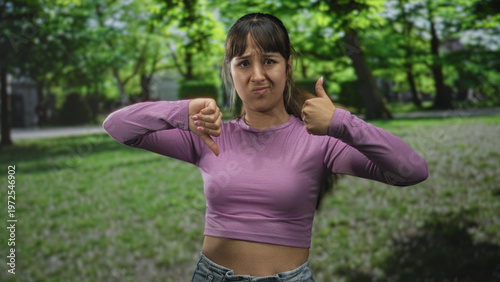 Fotografi Young woman showing thumbs up and thumbs down gesture in forest wearing purple crop top and expressive face; ambivalence mixed feelings