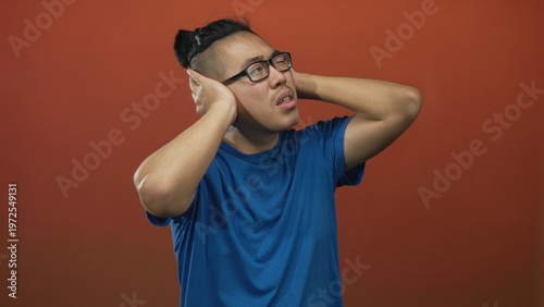 Foto Man holding hands over ears and wincing in a studio with a warm orange backdrop and blue shirt; discomfort