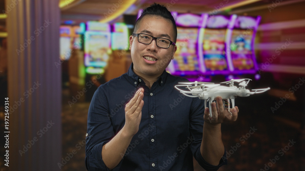 Fototapeta premium Man holding white quadcopter drone in one hand and gesturing with other while speaking in casino building with slot machines visible; curiosity.