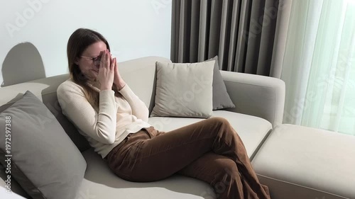 Woman sits on sofa at home holding head from fatigue. Female leans back touching temples with tired expression after long day. Condition suggests exhaustion headache and need for rest and