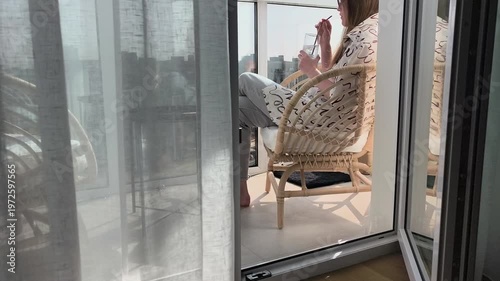 Woman relaxes on balcony in rattan chair enjoying quiet break. Female spends leisure time at home resting and feeling comfortable. Scene expresses balance between pause and everyday urban