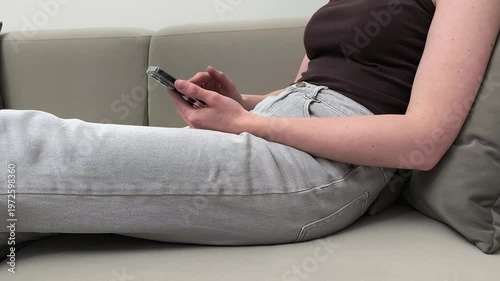 Woman sits on sofa and scrolls social media feed on smartphone at home. Female checks news content through mobile device using fingers. Concept online browsing, digital lifestyle, leisure