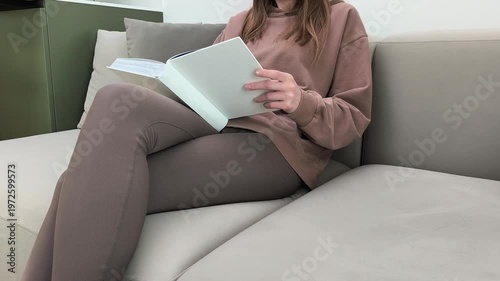 Woman turns book pages while resting on sofa indoors. Female flips sheets reading content during calm home time. Learning habit and relaxed lifestyle concept in apartment living room..
