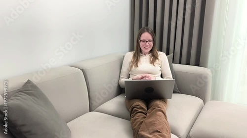 Woman lies on sofa holding laptop and laughs during video call at home. Female in glasses enjoys online conversation smiling through remote communication. Lifestyle connection concept for