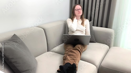 Laptop work continues as woman on sofa touches neck feeling pain. Female experiences discomfort from bad posture during remote job at home. Health concept related to sedentary lifestyle and