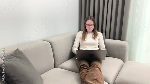 Woman sits on sofa stretching legs and picks laptop to start remote job. Female prepares device initiating online work activity from home. Business freelance concept with digital workspace