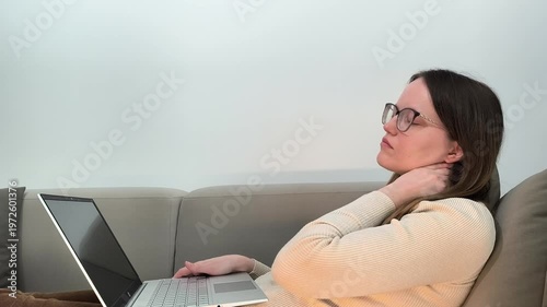 Laptop work continues as woman on sofa touches neck feeling pain from posture. Female experiences discomfort during remote job at home due to bad position. Health concept linked with